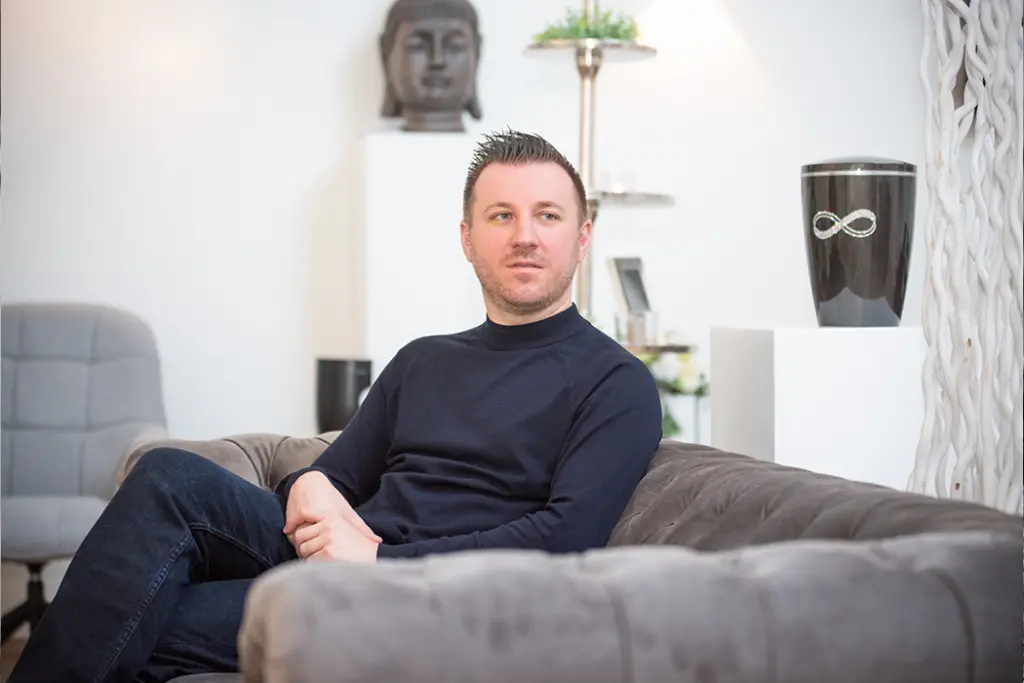 Trauerredner Torsten Schröder auf einer Couch sitzend, im Hintergrund steht eine Urne